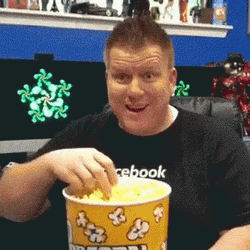 En person med kort hår sitter vid ett skrivbord, ler och äter popcorn från en stor gul hink i ett rum med skärmar och hyllor i bakgrunden.