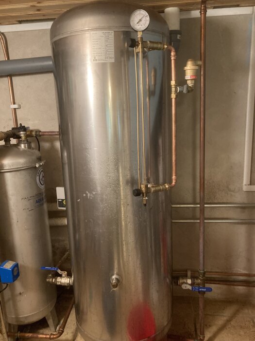 Hydrofor med anslutna kopparrör och en tryckmätare ovanpå, omgivet av ett annat grått metalliskt kärl och rörmotvikter.