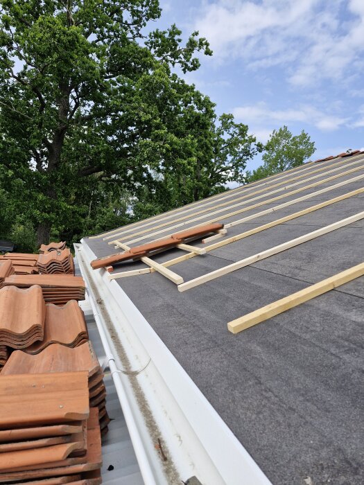 Takbyte med takpannor, strö- och bärläkt synliga på taket, delvis upplyfta pannor vid fotrännan, träd i bakgrunden.