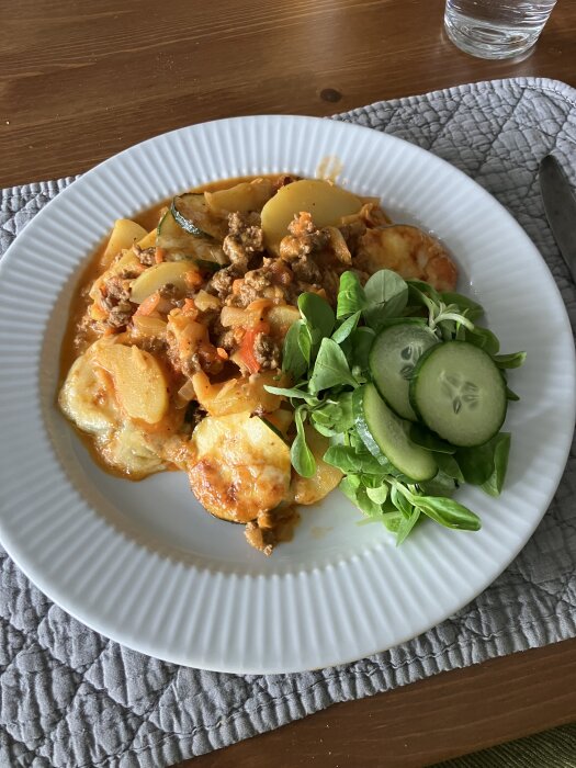 Tallrik med moussaka, innehållande potatis, zucchini och köttfärssås med grönsaker, serverad med en sallad av maché och gurkskivor.