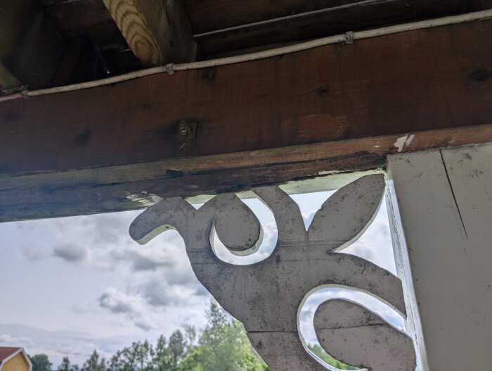 Rötangripen balk med extra bultad balk och dekorativt trästöd syns under takfoten i ett hus, med träd och molnig himmel i bakgrunden.