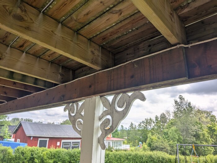Närbild på en träbalk under en altan med en extra bultad balk. Omgivningen visar gröna buskar och ett rött hus i bakgrunden.