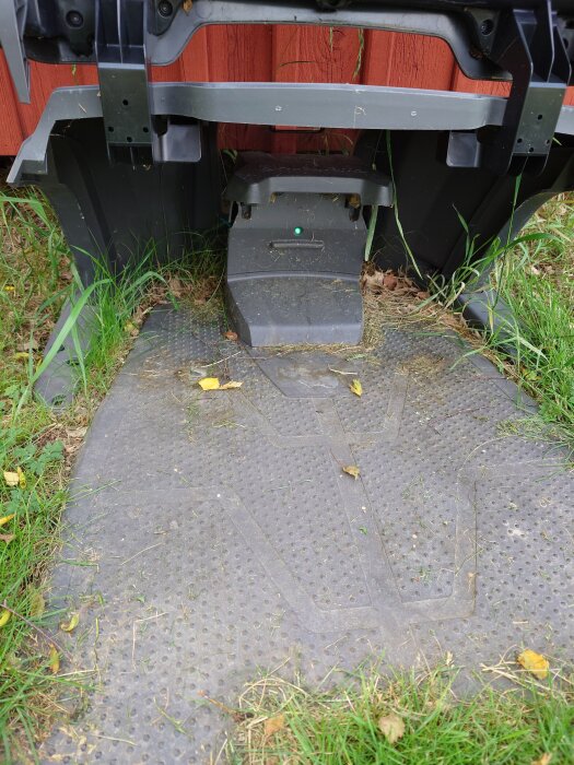 Laddstation för robotgräsklippare vid rött träskjul, delvis överväxt av gräs.