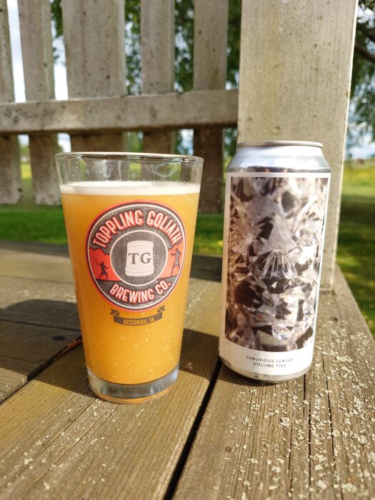 Ett glas fyllt med gulaktig öl från Toppling Goliath Brewing Co. bredvid en burk på ett träbord utomhus.
