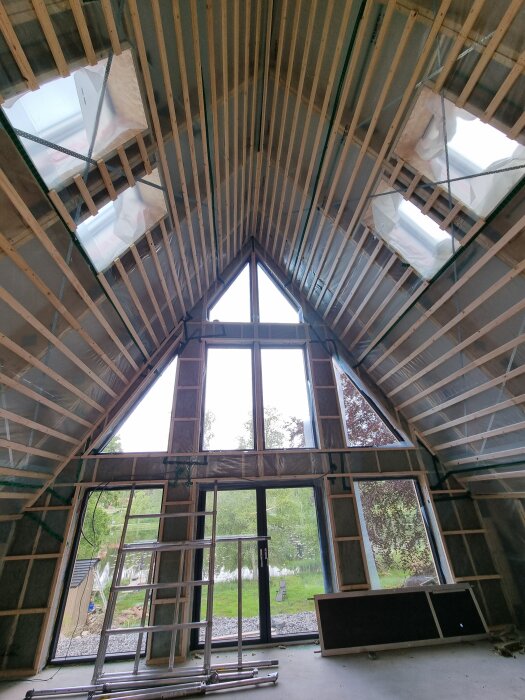 Byggarbetsplats under renovering med takbjälkar och stora fönster mot trädgården. Stege och byggmaterial vid glasdörr.