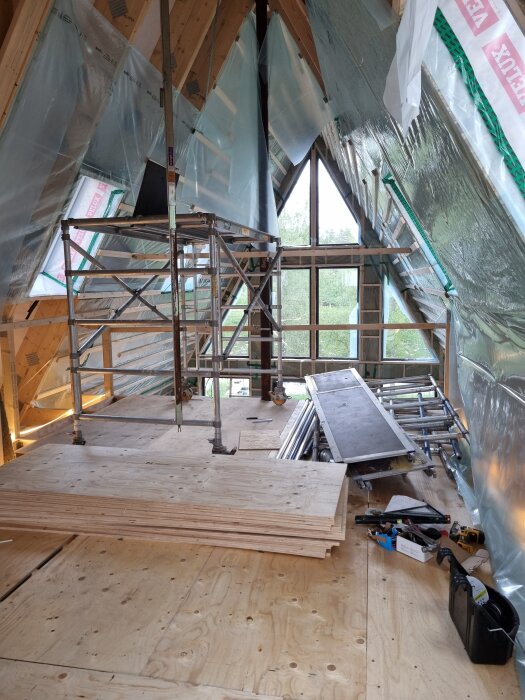 Byggarbetsplats i högdel med ställning och plast, brädor staplade på trägolv. Inre takbeklädnad av plast syns, verktyg och material vid sidan.