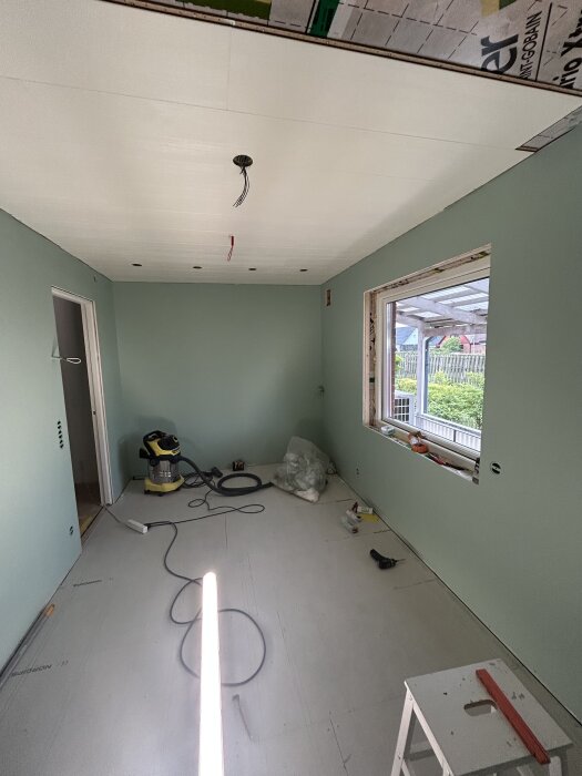 Rum under renovering med nymålade gröna väggar, plastat tak, och olika verktyg samt material på golvet. Ett fönster ger utsikt över en trädgård.