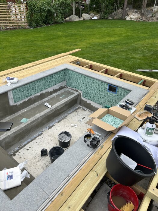 En pågående poolkonstruktion omgiven av grön gräsmatta, visar påbörjad plattsättning och byggmaterial som hinkar och plattor runt om.