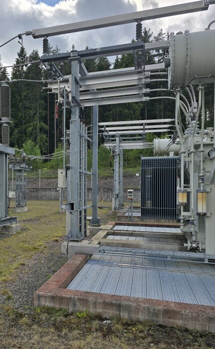 Utomhusanläggning med transformator och ställverk med isolatorer och ledningar, omgiven av skog i bakgrunden.