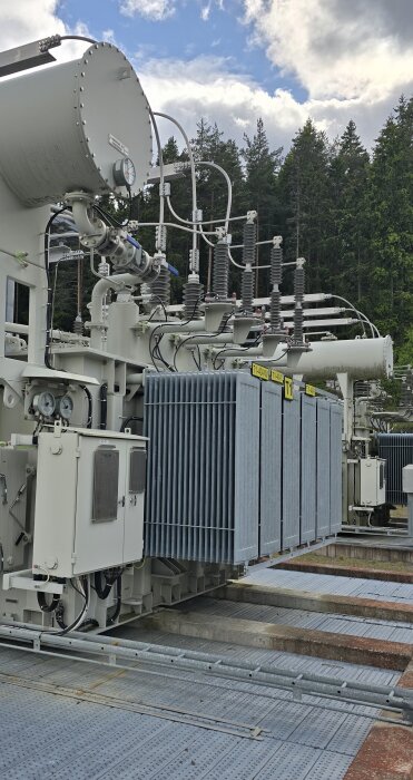 Transformator med rör och isolatorer på en metallplatta, omgiven av tallskog och blå himmel.