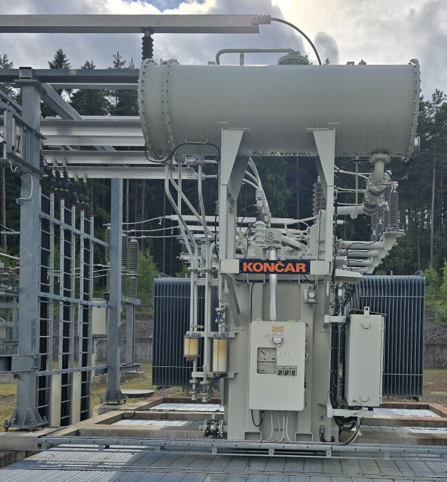 Transformator utomhus med anslutningar, avledare och skenor. Märkt med "Končar". Omgiven av skog. Sesskydd, mätutrustning och kylsystem synliga.