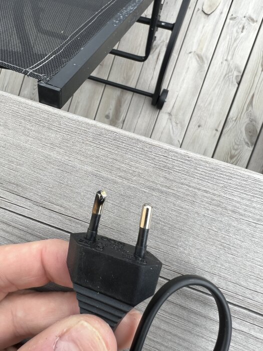Skadad elkontakt med brännmärke på ena stiftet, hålls i handen över en träterrass.