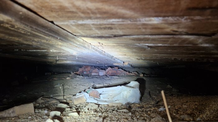 Bild av en trång krypgrund med synliga träbjälkar i taket, tegelbitar och diverse skräp på marken.