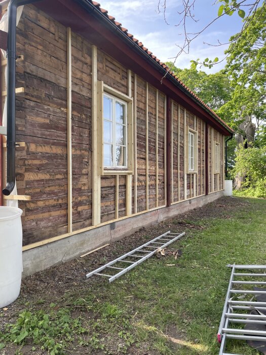 Vägg under panelbyte på hus med synligt trä, ställning på marken och grönska i bakgrunden.