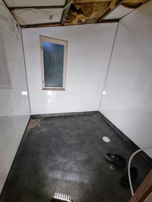 Ett badrum med vita våtrumsskivor och svart golv, ett litet fönster på väggen, skador i taket och vattensäkert skydd saknas vid hörn.