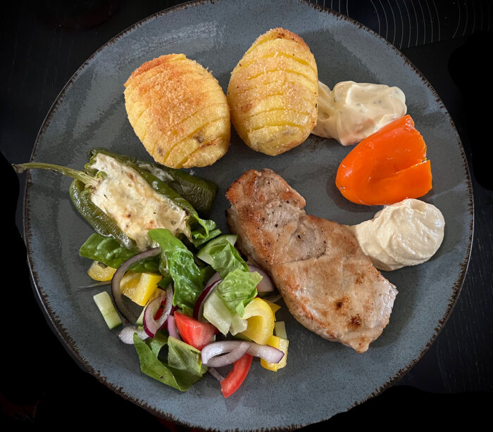 Tallrik med fläskkotlett, hasselbackspotatis, sallad, fylld chili, hummus och en klick bearnaisesås.