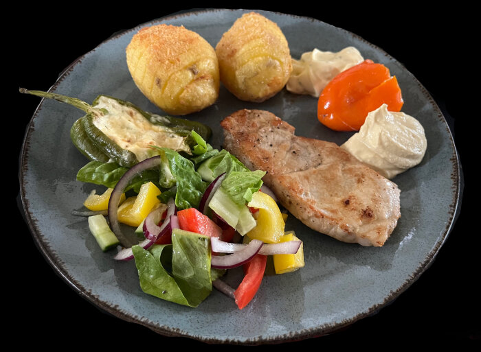 Tallrik med fläskkotlett, hasselbackspotatis, sallad, fylld chili, hummus och bearnaisesås.