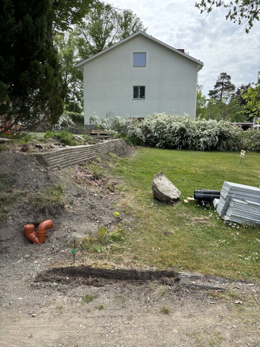 Gräsmatta med jordhög på sidan, orange avloppsrör, gräsmatta och träd i bakgrunden. Hus i bakgrunden.