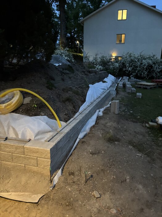 Mur byggd av betongblock, omgiven av jord, med ett hus och grönska i bakgrunden, under skymningen.