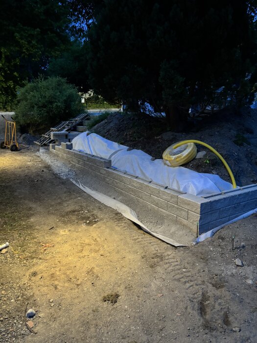 Mur av cementblock under byggnation, med jord och växtlighet i bakgrunden, synligt rör och vit duk på marken.