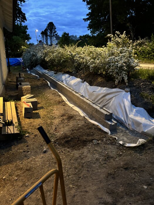 Murprojeket under arbete med staplade betongblock längs en blomsterrabatt, kartongbitar och verktyg synliga på marken.