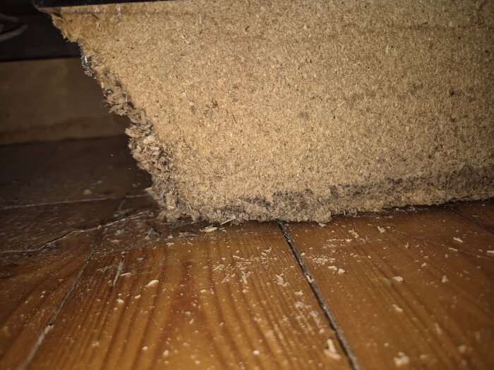 Närbild på undersidan av en kökssockel med fuktskador och mögel på trä och runt stommaterial, stående på ett furugolv.