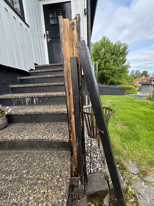 Trappräckesstolpe med ruttnat trä runt en stålstolpe vid en utomhustrapp; omgivningarna inkluderar gräsmatta och husfasad.