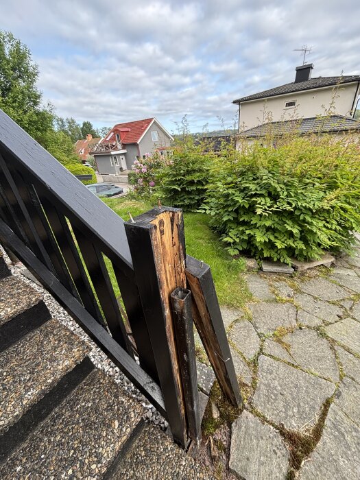 Trästolpe vid trappan med rutten kärna, exponerad stålstolpe och omgivande gröna buskar samt stenlagd gång.