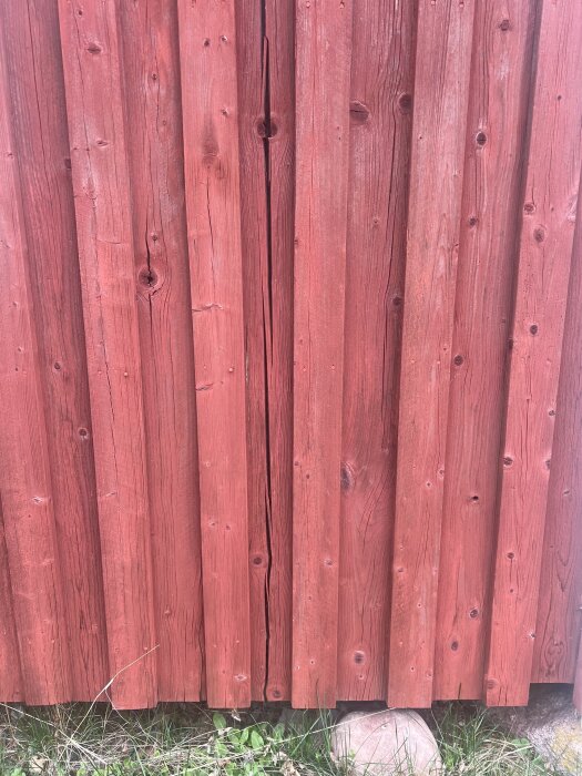 Röd lockpanel på ett gammalt hus med spruckna brädor i behov av reparation och målning. Grästrån syns vid panelens bas.