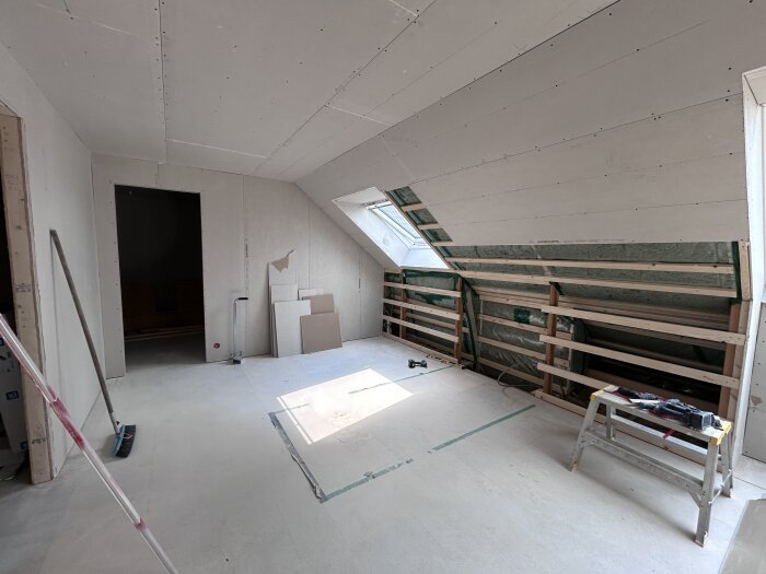 Rum under renovering med gipsskivor på väggar och tak, takfönster, träreglar och verktyg. Städutrustning och byggmaterial syns på golvet.