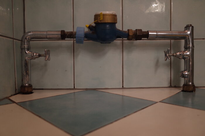 Vattenmätare installerad med Securex-kopplingar och kopparrör mot kaklad vägg och golv i äldre hus från 1962.