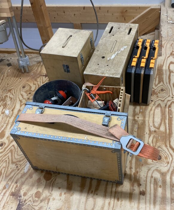 Plywoodlådor, en hink med verktyg, och plastförvaringslådor på ett trägolv i en byggmiljö.
