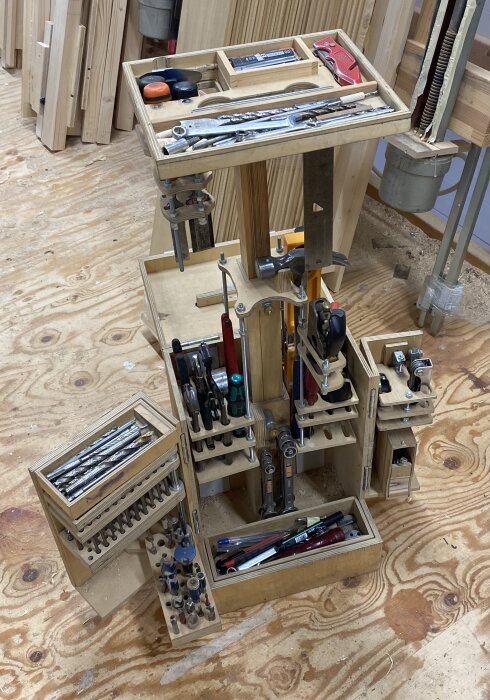 Plyfalåda utformad som verktygshållare, fylld med diverse handverktyg och borr, placerad på ett träbordsunderlag i verkstadsrum.