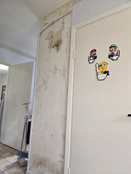 Vägg i hall med borttagen vävtapet, visar skadade och fläckiga ytor. Dörr dekorerad med pixelinspirerade figurer.