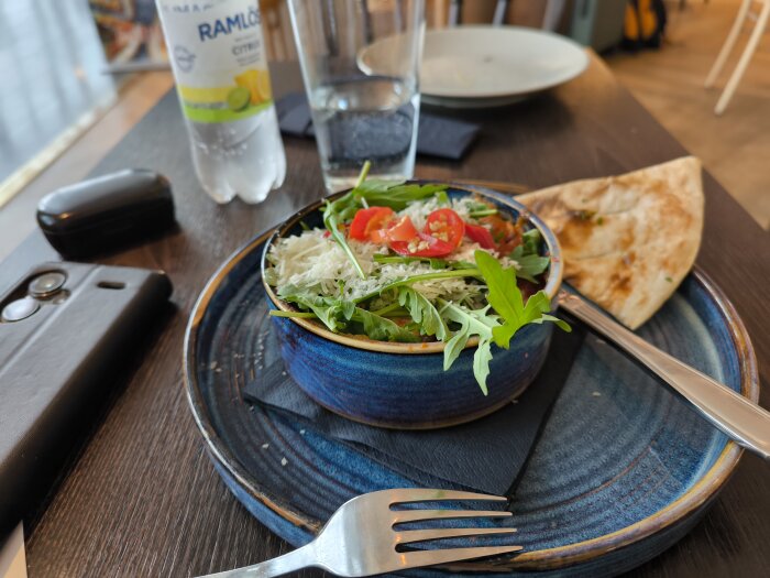 Lasagne med ruccola och tomater serverad i en blå skål på en svart tallrik, bredvid vitlöksbröd och en flaska Ramlösa citron.