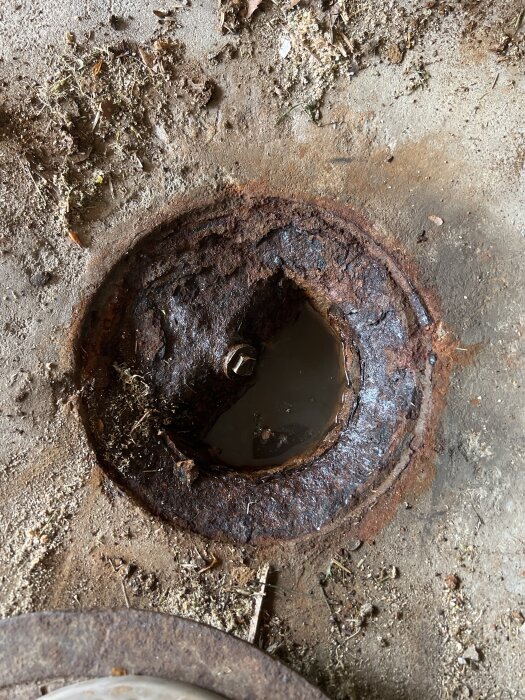 Rostig avloppsbrunn i garagegolv från 70-talet, delvis fylld med vatten och omgiven av smuts och skräp.