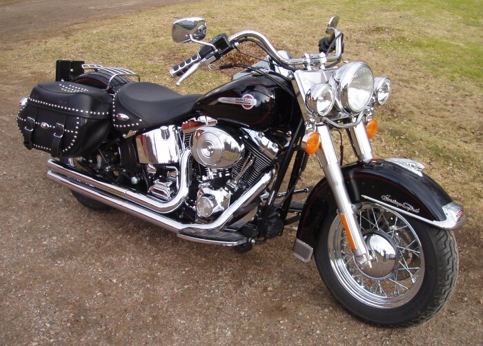Svart motorcykel med kromdetaljer, lädersadlar och fotplattor, parkerad på en grusväg. Ingen vindruta synlig.