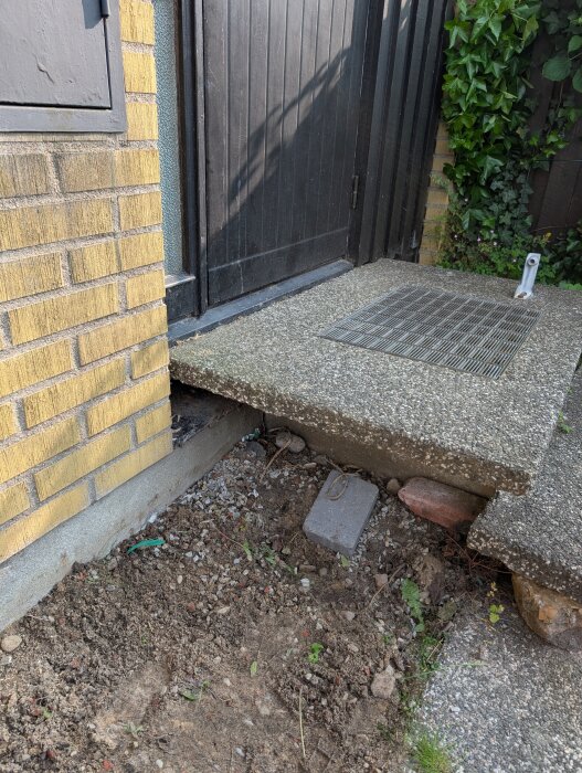 Kuvan keskellä on betoninen sisäänkäyntiaskelma tiiliseinän vieressä, kasvillisuutta ja paljastettua maata näkyy alapuolella.