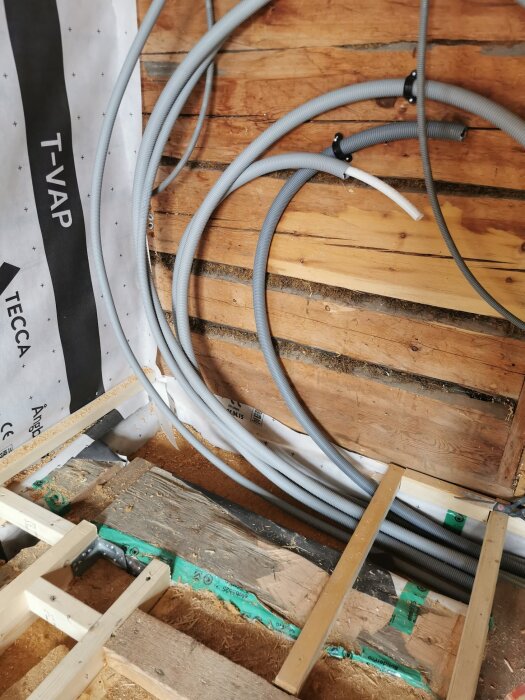 Gråa rör med böjar monterade på en trävägg i ett rum under renovering, med isolering och träreglar på golvet.