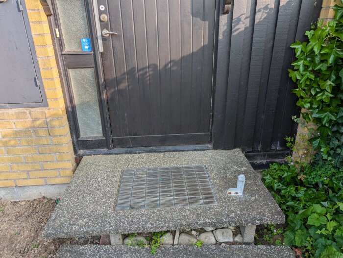 Mustan ulko-oven edessä betoninen porras askelmilla ja metalliritilä keskellä, vieressä keltatiilinen ja laudoitettu seinä.