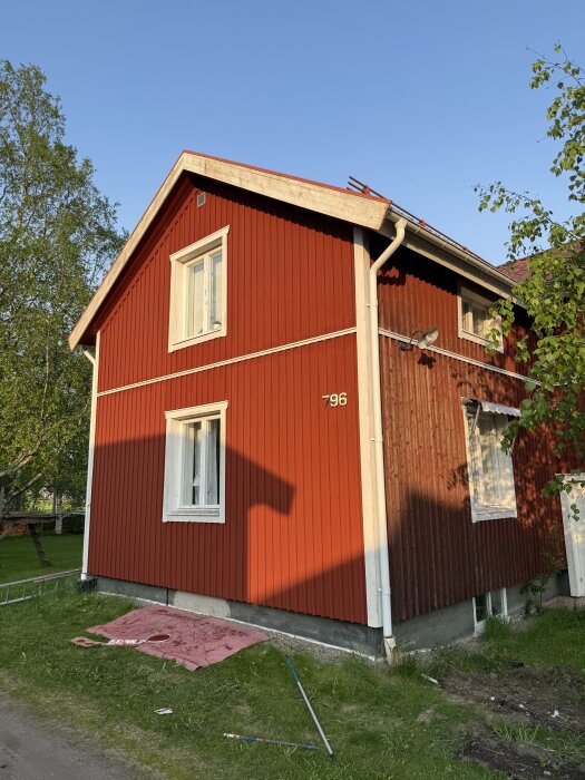 Röd husfasad till hälften nymålad, solbelyst. Stege och målarredskap på marken.