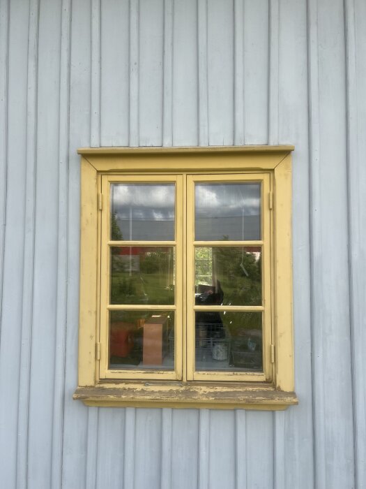Gult träfönster på grå träpanel, med spröjs och karm i herrgårdsgul och ockragul nyans, delvis sliten färg.