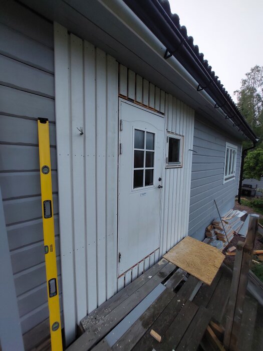Yttervägg med nästan färdig lockpanel, vit dörr och fönster. En gul vattenpass står lutad mot väggen.