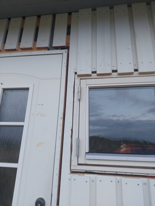 Närbild på vit lockpanel under konstruktion vid dörr och fönster. Panelen saknar foder och det syns skruvar förmonterade.