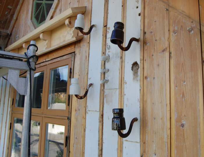 Rustik träfasad med fyra isolatorer för luftledning monterade, två vita och två svarta, samt ett fönster och en snedställd bräda ovanför.