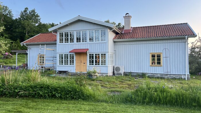 Blågrått trähus med röda takpannor; en gul dörr; fönster med vita spröjs och det ena med gult foder; tre ställningar i trädgården.