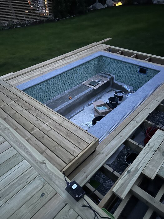 En underbyggd pool med trätrall runt, täckt med små gröna plattor och omgivande verktyg och material.