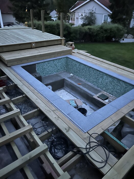 Byggarbetsplats med en oavslutad pool under tak. Trädäck omger poolen, verktyg och material synliga runt om.