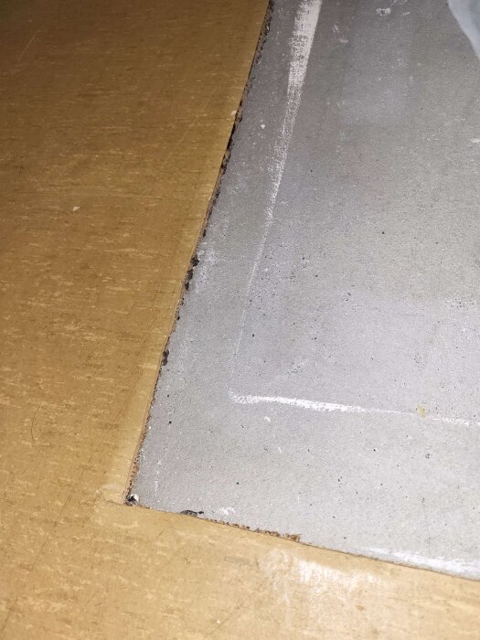 Bild på ett grått golv med en sliten kant där materialet ser ut att flagna. En del av ett mörkt nät syns längs kanten. Golvet verkar vara linoleum eller liknande.