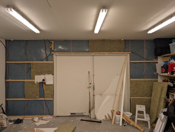 Garaget inifrån, visar delvis rivna väggar runt existerande garageport. Isolering och träreglar synliga, nedre delen av väggen är riven och byggmaterial ligger på golvet.